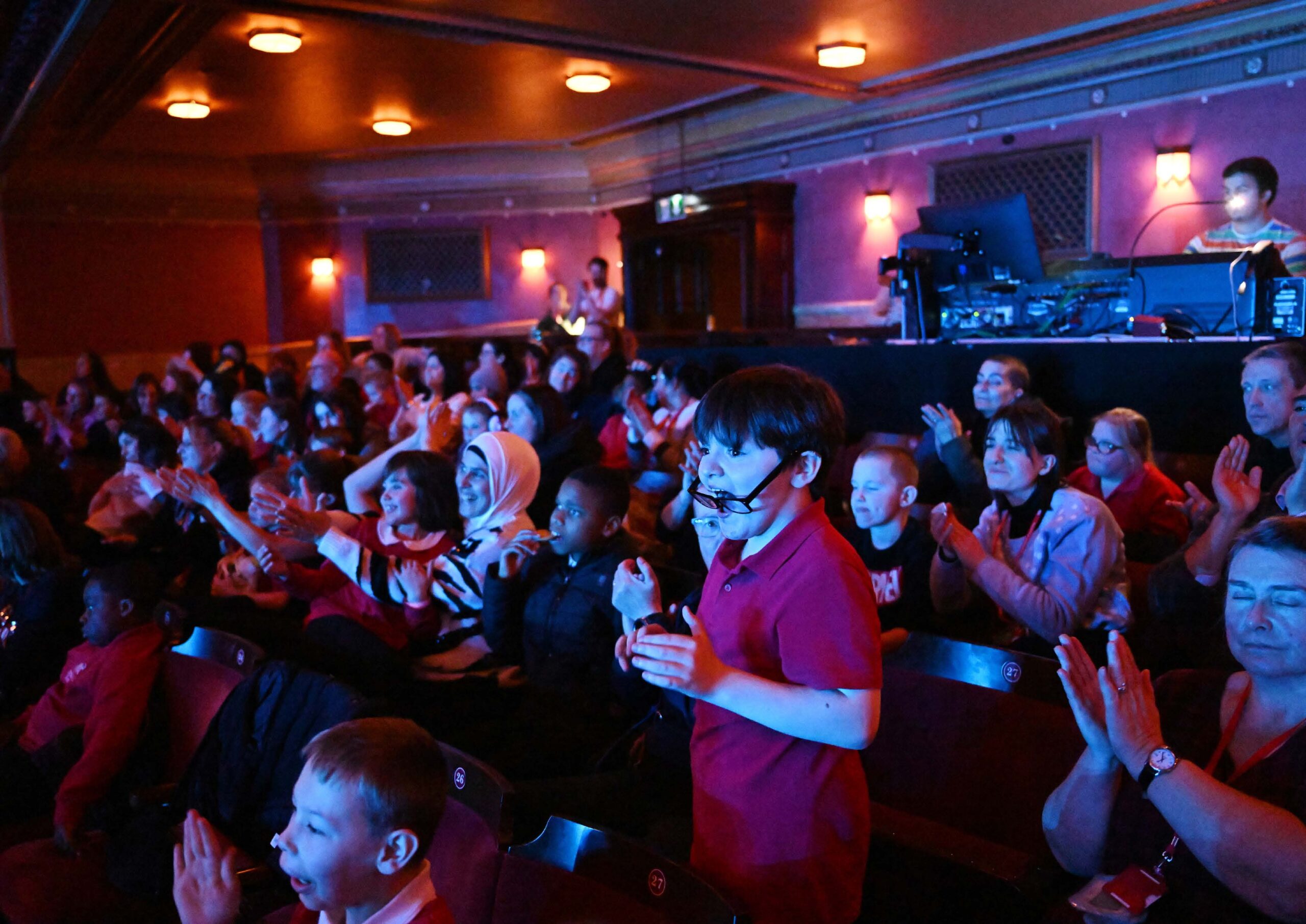 Children and adults sit in theatre seats, some clapping and smiling, while others watch the stage; staff are seen at a control booth in the background.