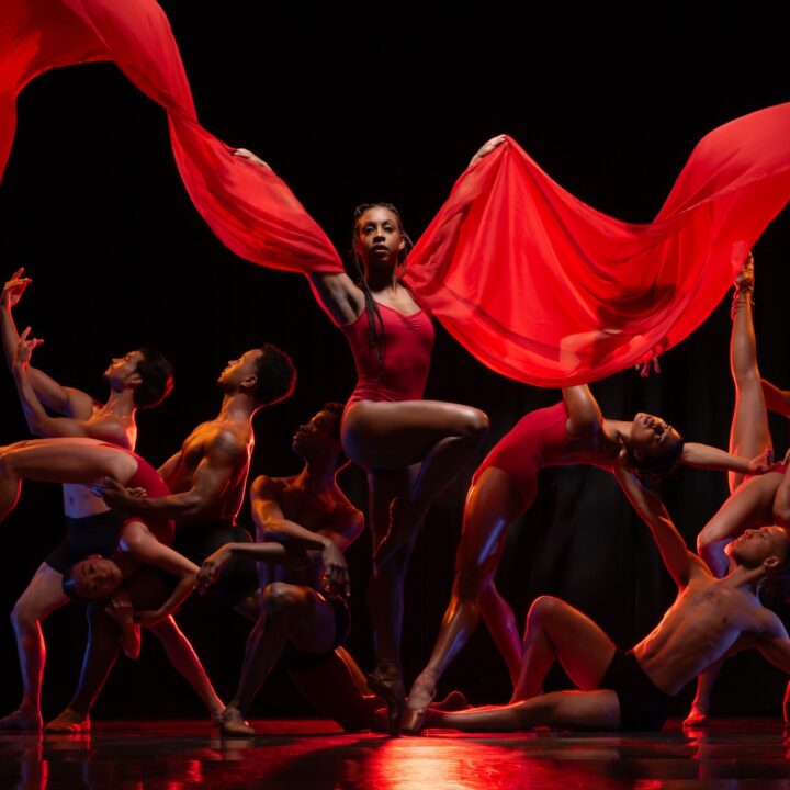 A group of dancers pose dramatically on stage, with one dancer in the center holding flowing red fabric above her head.