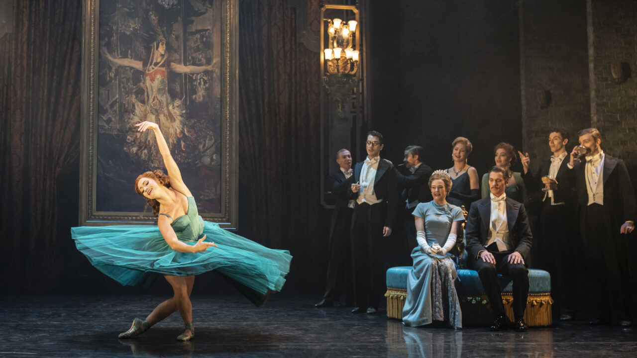 A ballerina in a blue dress performs on stage in front of a painting, while formally dressed people watch from the side and a chandelier hangs overhead.