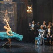 A ballerina in a blue dress performs on stage in front of a painting, while formally dressed people watch from the side and a chandelier hangs overhead.