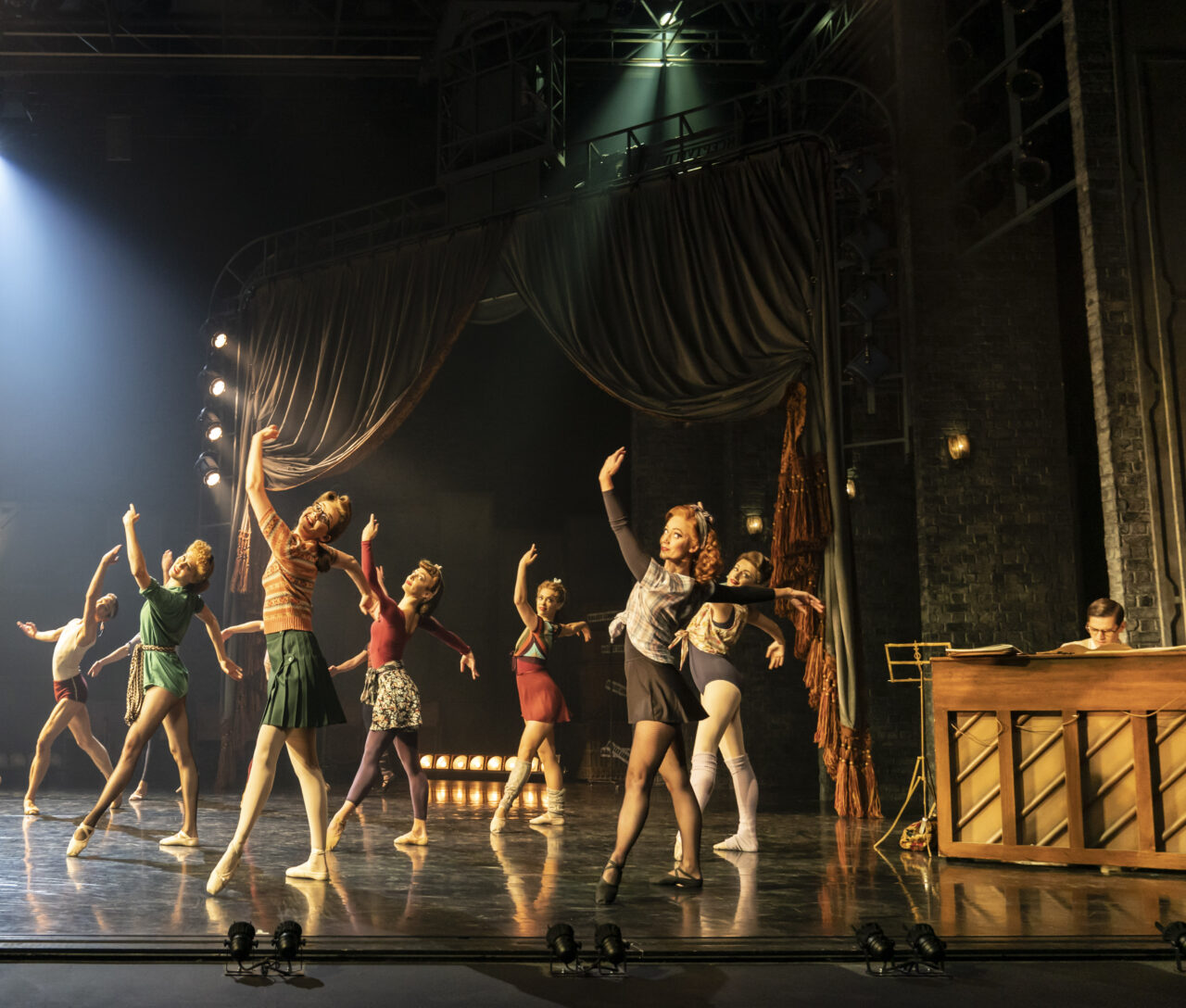 A group of dancers rehearses on stage in front of a pianist, with theatrical lighting and curtains in the background.