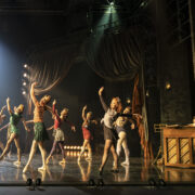 A group of dancers rehearses on stage in front of a pianist, with theatrical lighting and curtains in the background.