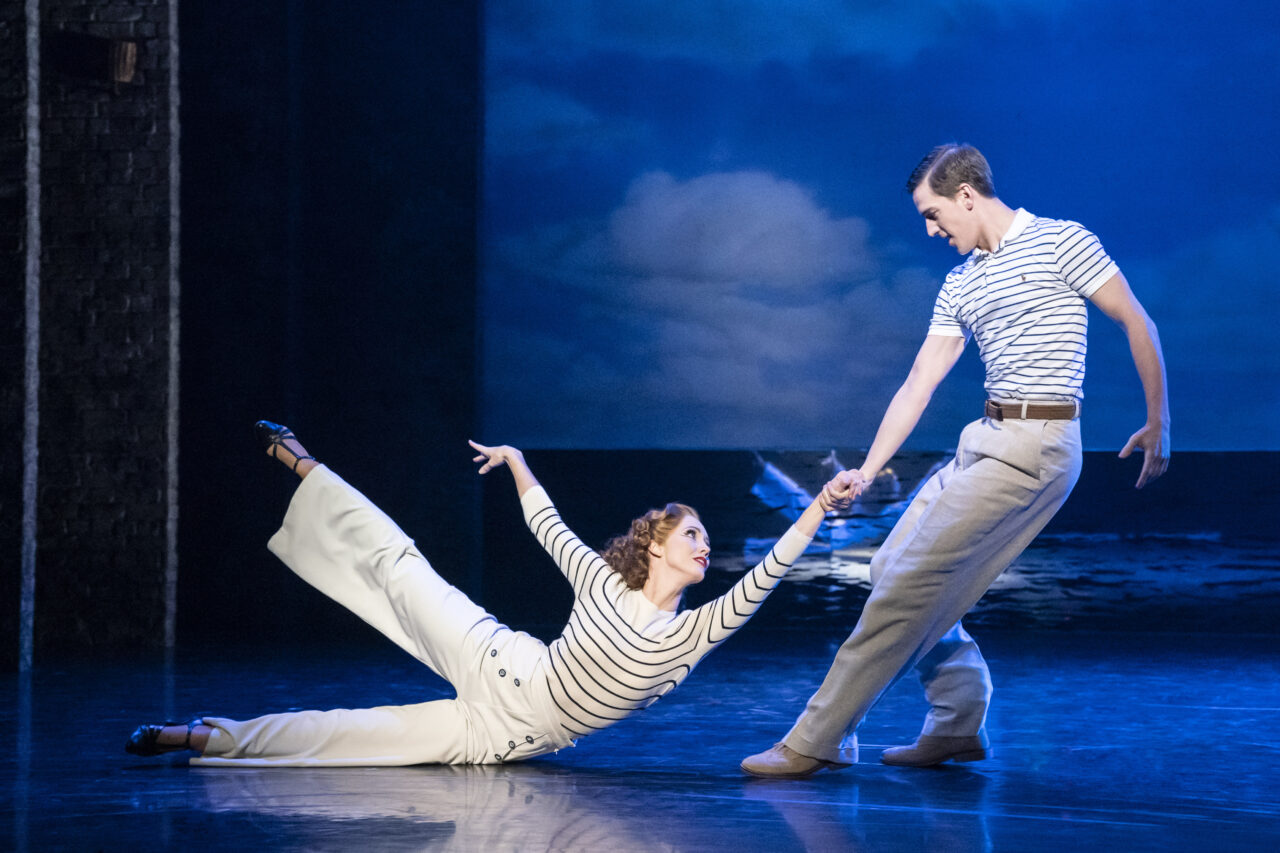 A woman in white pants and a striped shirt poses on the floor, holding hands with a standing man in similar attire, performing on stage with a seascape backdrop.