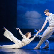 A woman in white pants and a striped shirt poses on the floor, holding hands with a standing man in similar attire, performing on stage with a seascape backdrop.