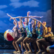 A group of dancers in vintage swimsuits pose together on stage with beach balls, against a backdrop of blue sky and ocean.