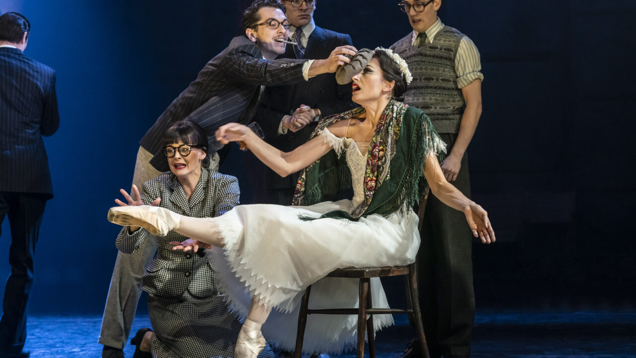 A group of actors in vintage costumes surround a seated ballerina on stage; one man reaches toward her head as she balances en pointe on one foot.