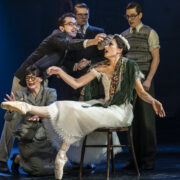 A group of actors in vintage costumes surround a seated ballerina on stage; one man reaches toward her head as she balances en pointe on one foot.