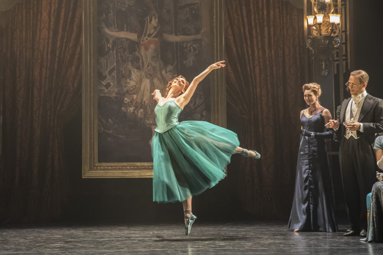 A ballerina in a teal dress performs on stage in front of a large painting, while three formally dressed people watch from the side.