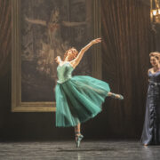 A ballerina in a teal dress performs on stage in front of a large painting, while three formally dressed people watch from the side.
