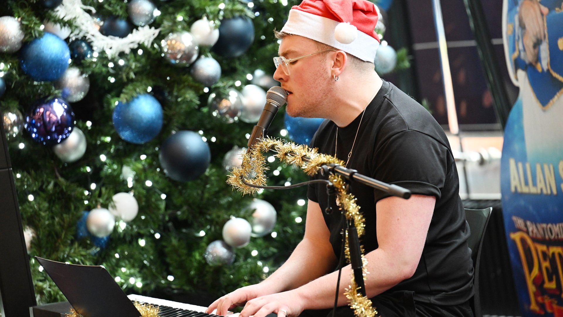 Person in a Santa hat plays keyboard and sings, with a decorated Christmas tree and festive decorations in the background.