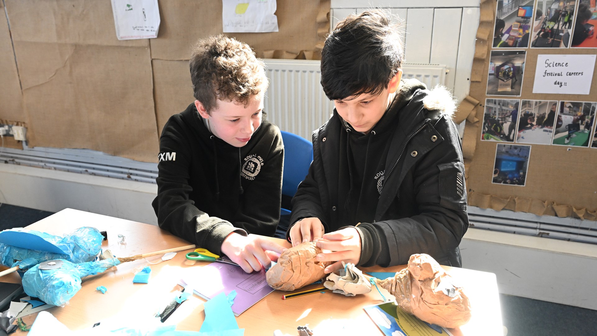 Two boys sit at a table in a classroom, working together on a papier-mâché model with craft materials scattered around them.