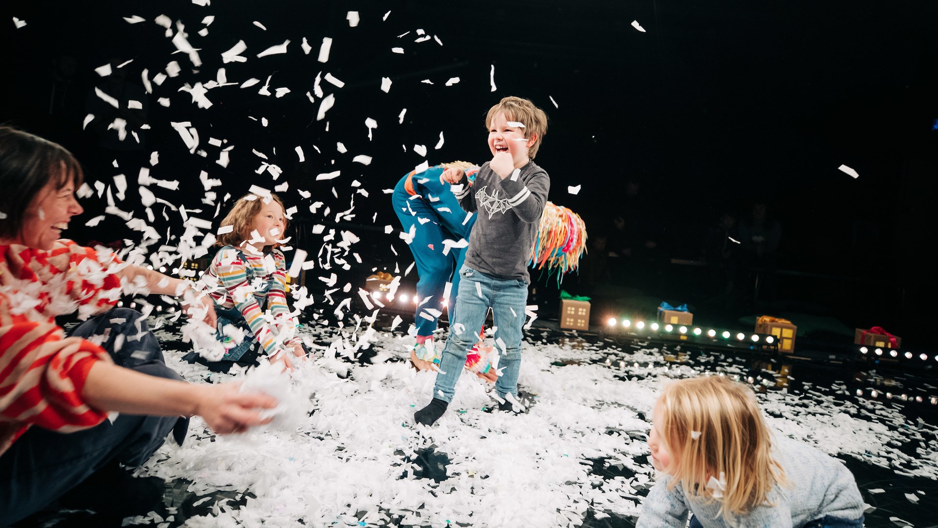 Children play joyfully amid scattered white confetti on a stage, with adults joining in and colorful houses visible in the background.