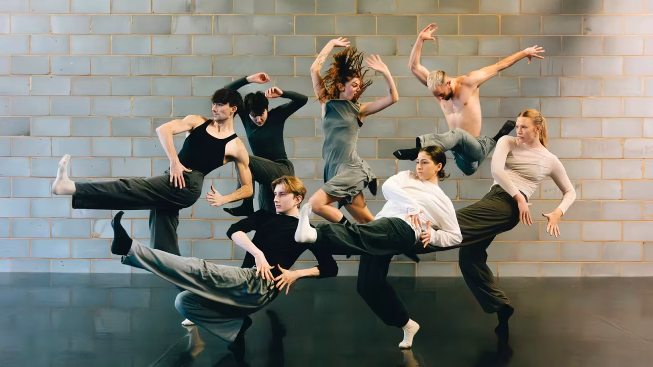 A group of six contemporary dancers in motion, exploring new horizons against a brick wall.