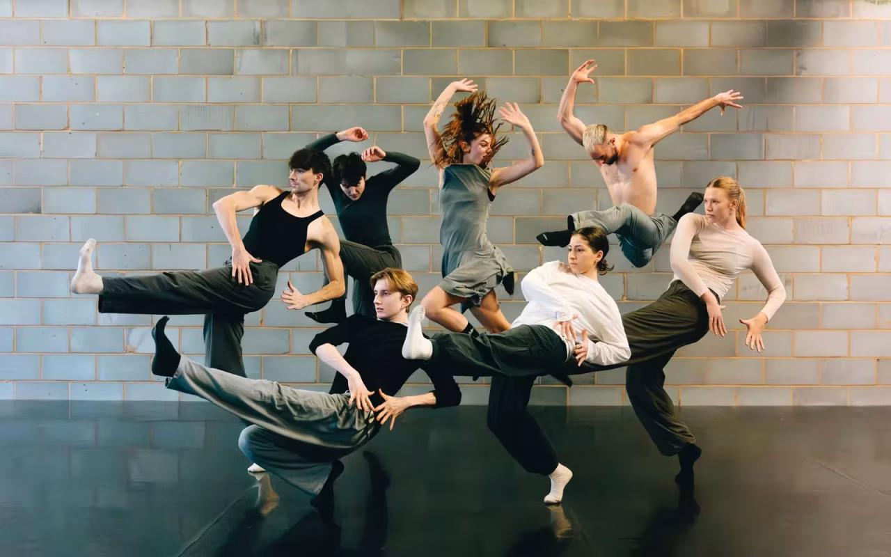 A group of six contemporary dancers in motion, exploring new horizons against a brick wall.