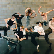 A group of six contemporary dancers in motion, exploring new horizons against a brick wall.